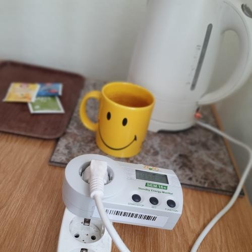 Im Vordergrund Strommessgerät, im Hintergrund Tasse, Wasserkocher und Teebeutel